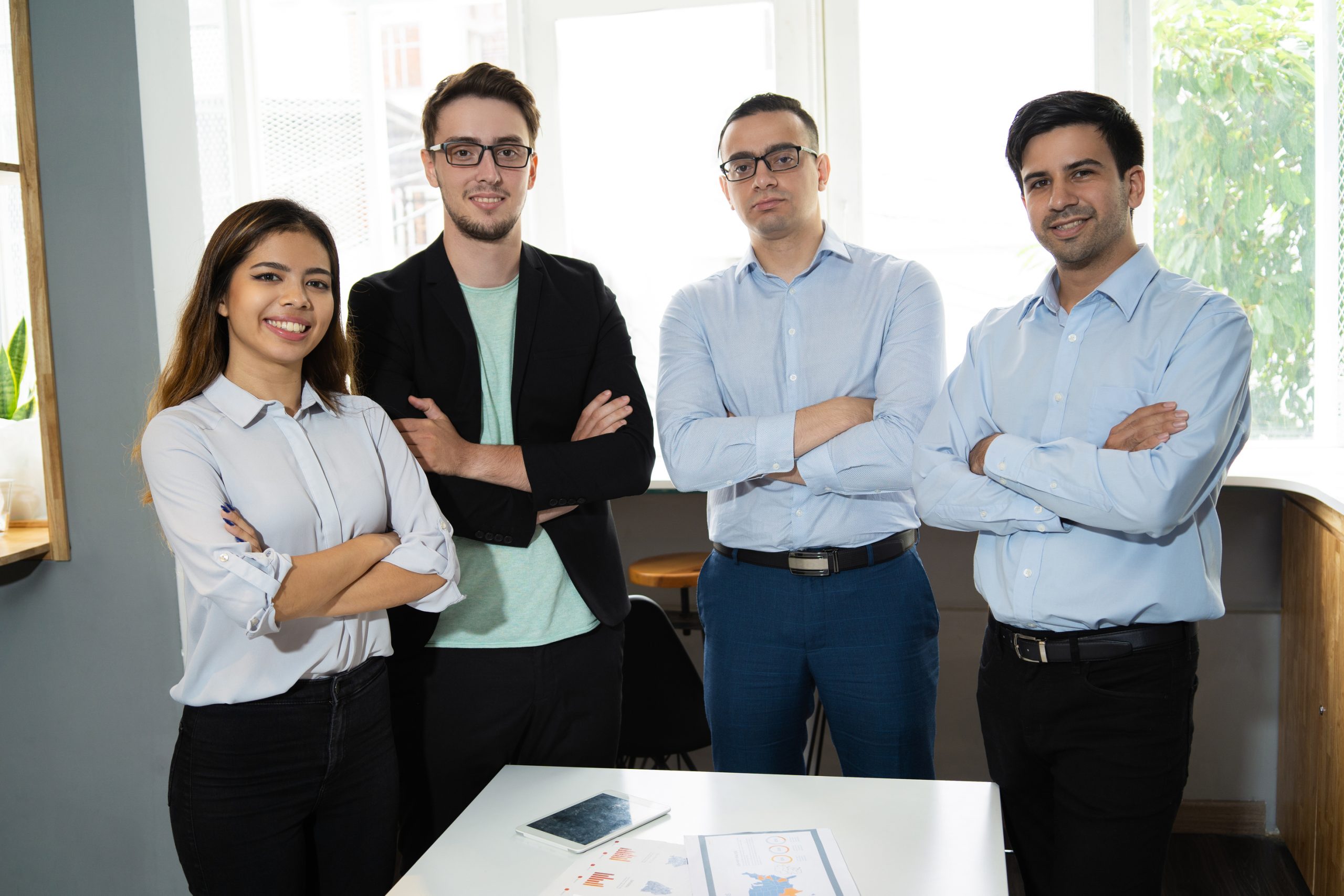 Positive successful business team posing at workplace. Group of four managers standing at meeting table with arms crossed. Successful teamwork concept