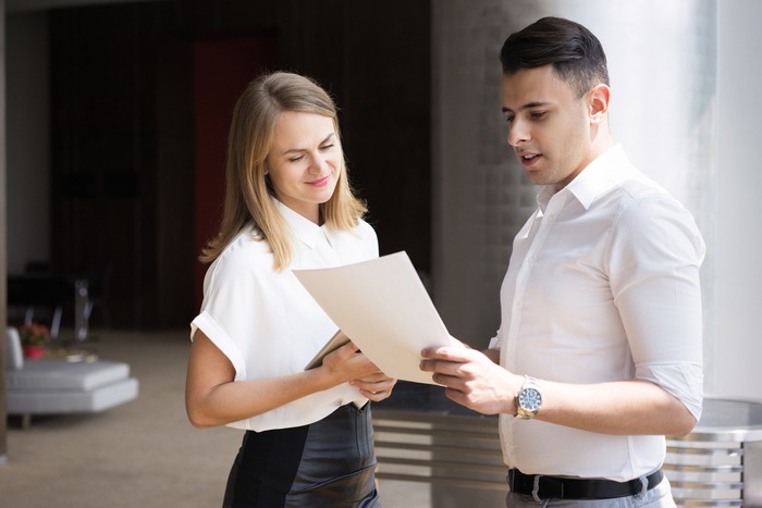 Young businessman reading document to smiling businesswoman. Caucasian colleagues examining document modern office. Partnership, cooperation concept