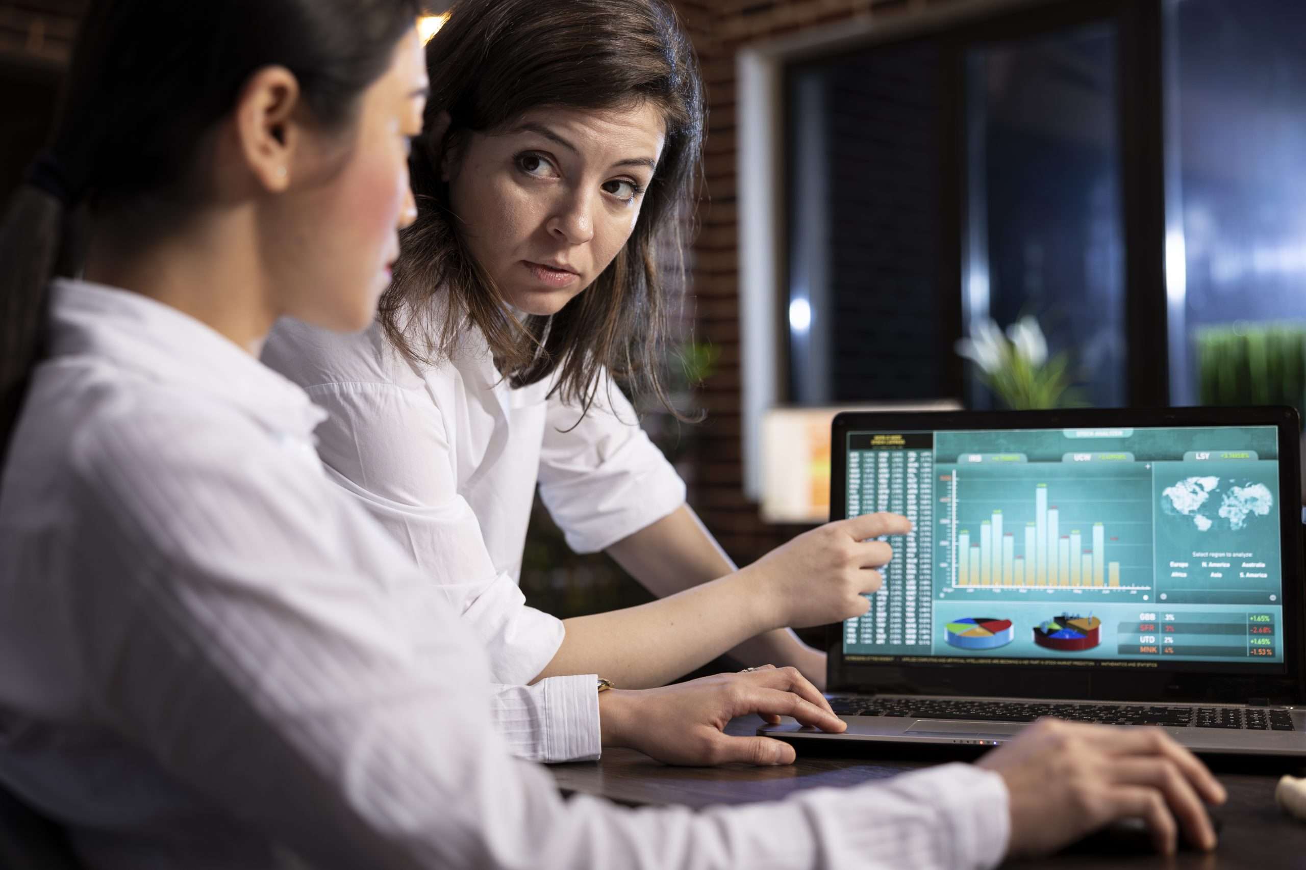 Two female colleagues collaborating on business analytics, reviewing data and discussing strategies on a laptop. They analyze financial information and plan tasks ahead in a professional workspace.