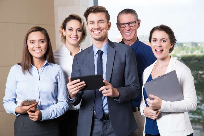 Group of five happy business people standing in conference room and smiling at camera. Young businessman holding tablet computer, businesswoman with clipboard, her Asian colleague with smartphone